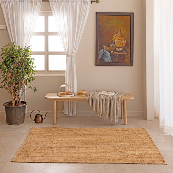 Handwoven natural jute area rug in warm beige and earthy tones, featuring a textured woven design styled in a cozy neutral room with a vase and book.