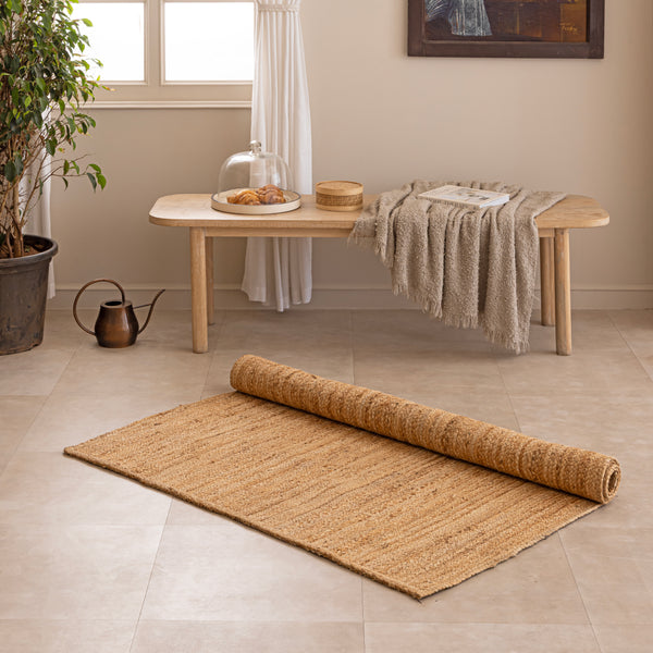 Handwoven natural jute area rug in warm beige and earthy tones, featuring a textured woven design styled in a cozy neutral room with a vase and book.