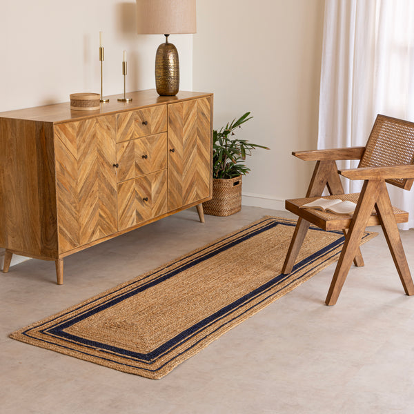 Handwoven natural jute runner rug with navy blue geometric border, styled in a warm modern living room with wooden furniture and a cane chair.