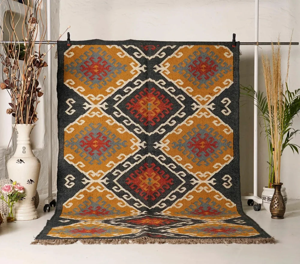 Large handwoven Kilim wool rug with a traditional geometric diamond motif in ochre yellow, charcoal black, terracotta rust, and grey, displayed vertically on a metal rack.