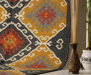 Large handwoven Kilim wool rug with a traditional geometric diamond motif in ochre yellow, charcoal black, terracotta rust, and grey, displayed vertically on a metal rack.
