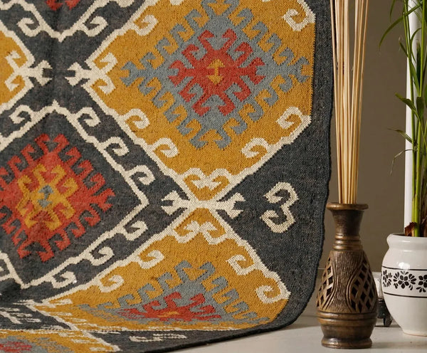 Large handwoven Kilim wool rug with a traditional geometric diamond motif in ochre yellow, charcoal black, terracotta rust, and grey, displayed vertically on a metal rack.